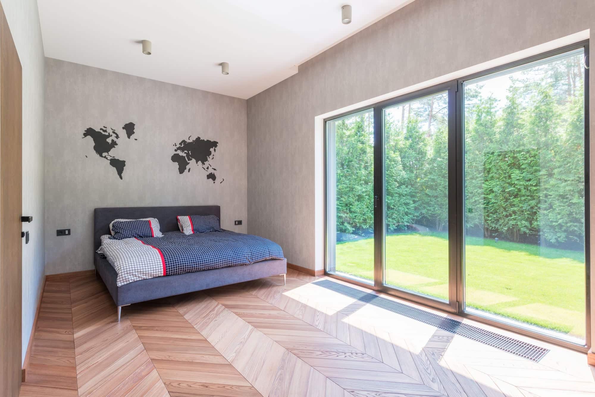 Chambre lumineuse au parquet en chevrons, avec une large baie coulissante à cadre gris donnant sur une pelouse