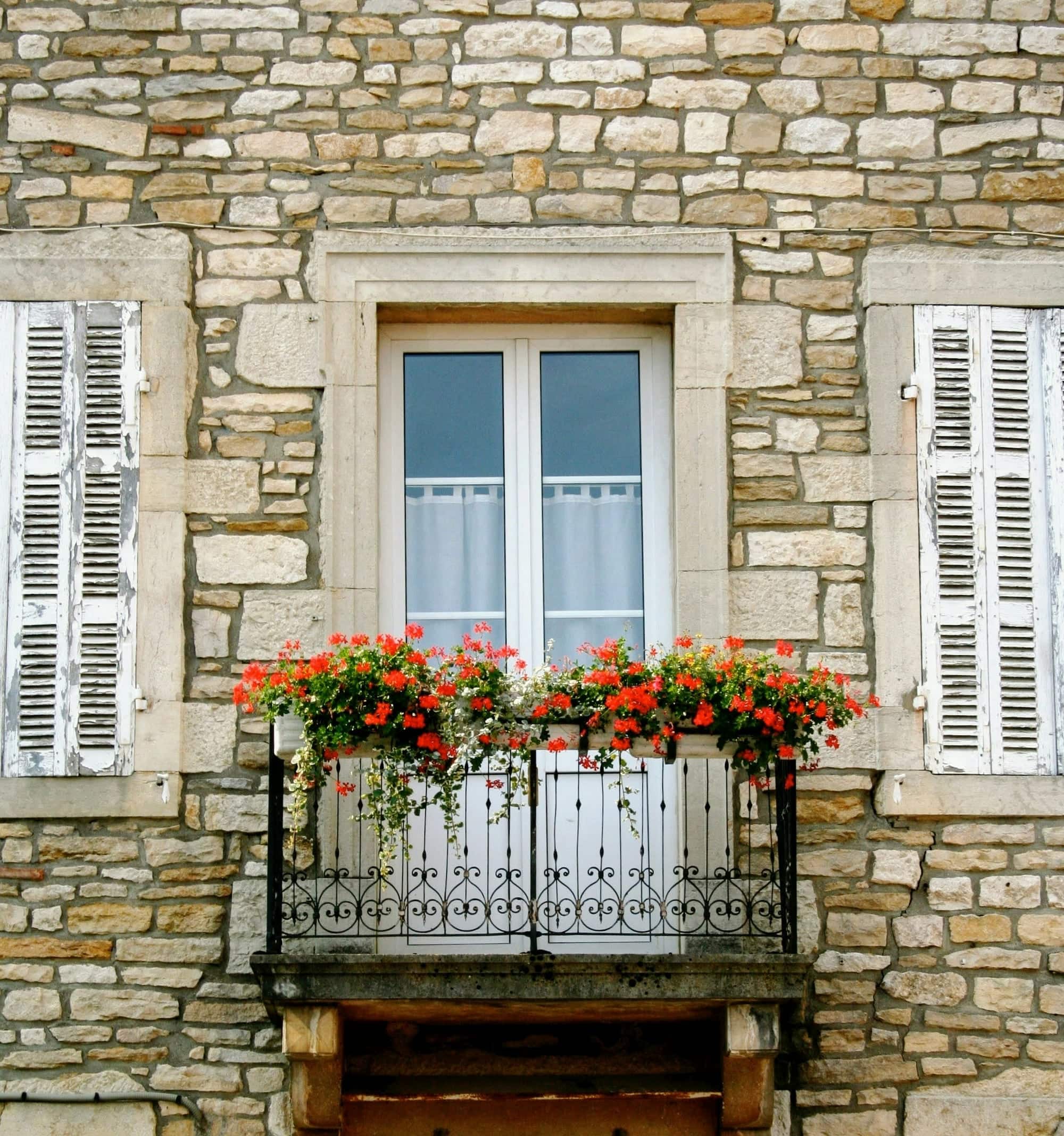 Façade en pierre avec une porte-fenêtre blanche encadrée de persiennes en bois et un balcon fleuri