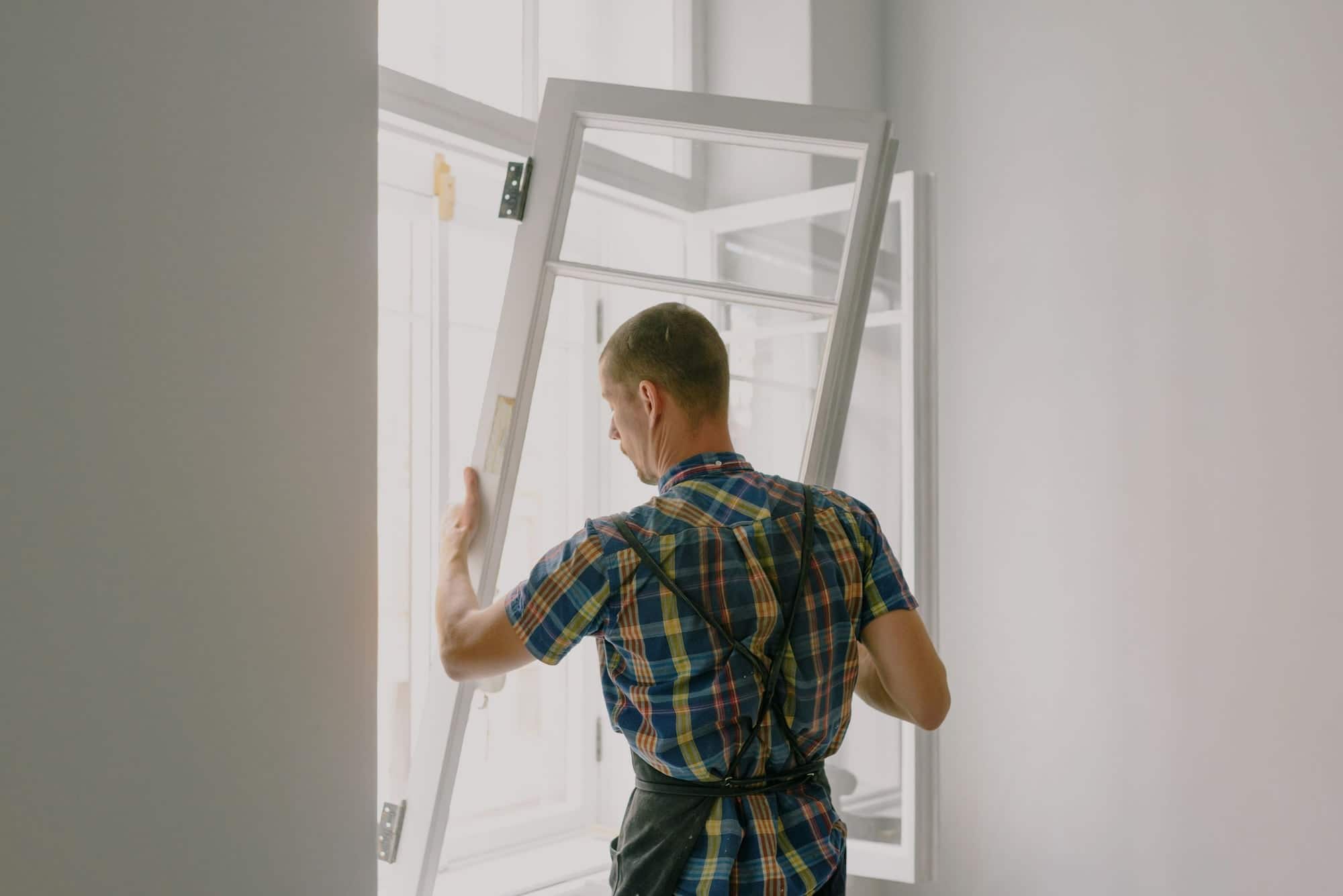 Poseur vu de dos mettant en place un châssis de fenêtre en bois blanc dans une pièce en rénovation